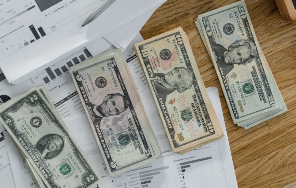 Stacks of US Dollar banknotes placed over financial documents and reports on a desk.